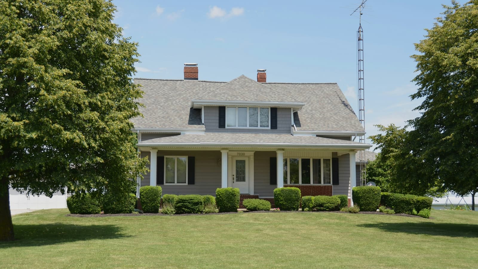 Two-story farmhouse with a gray asphalt-shingle roof, front porch, trimmed shrubs and a wide green lawn under a blue sky